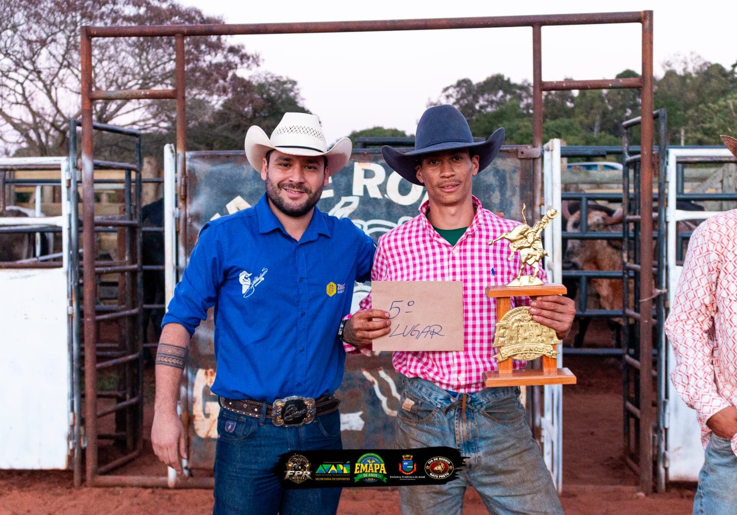 Marcos Ferraz aparece à direita, usando chapéu escuro e camisa xadrez rosa, segurando o troféu e uma placa com a indicação de 5º lugar. Ao lado dele, um integrante da organização posa para a foto. Ao fundo, aparecem a porteira e a estrutura da arena de rodeio.