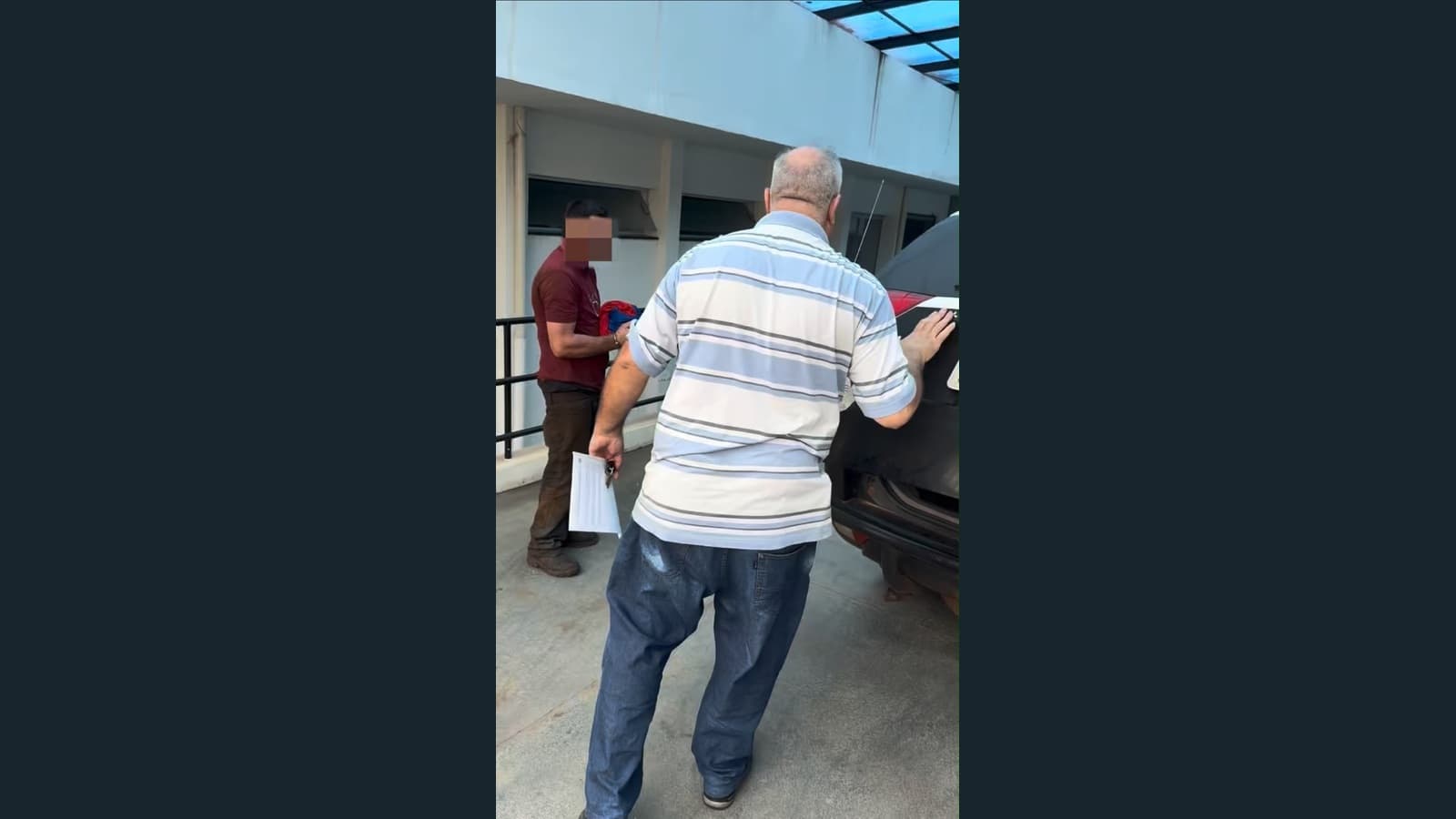 Fotografia mostra um homem de camiseta vinho com o rosto ocultado por pixelização, próximo a outro homem de costas, de camisa listrada, ao lado de um veículo. A imagem vertical foi mantida inteira, com faixas laterais escuras.